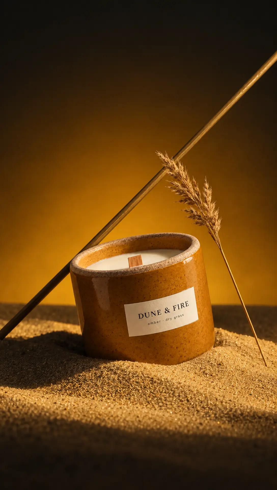 An ochre Dune & Fire candle in sand with dried grass, on an ochre-to-black gradient.
