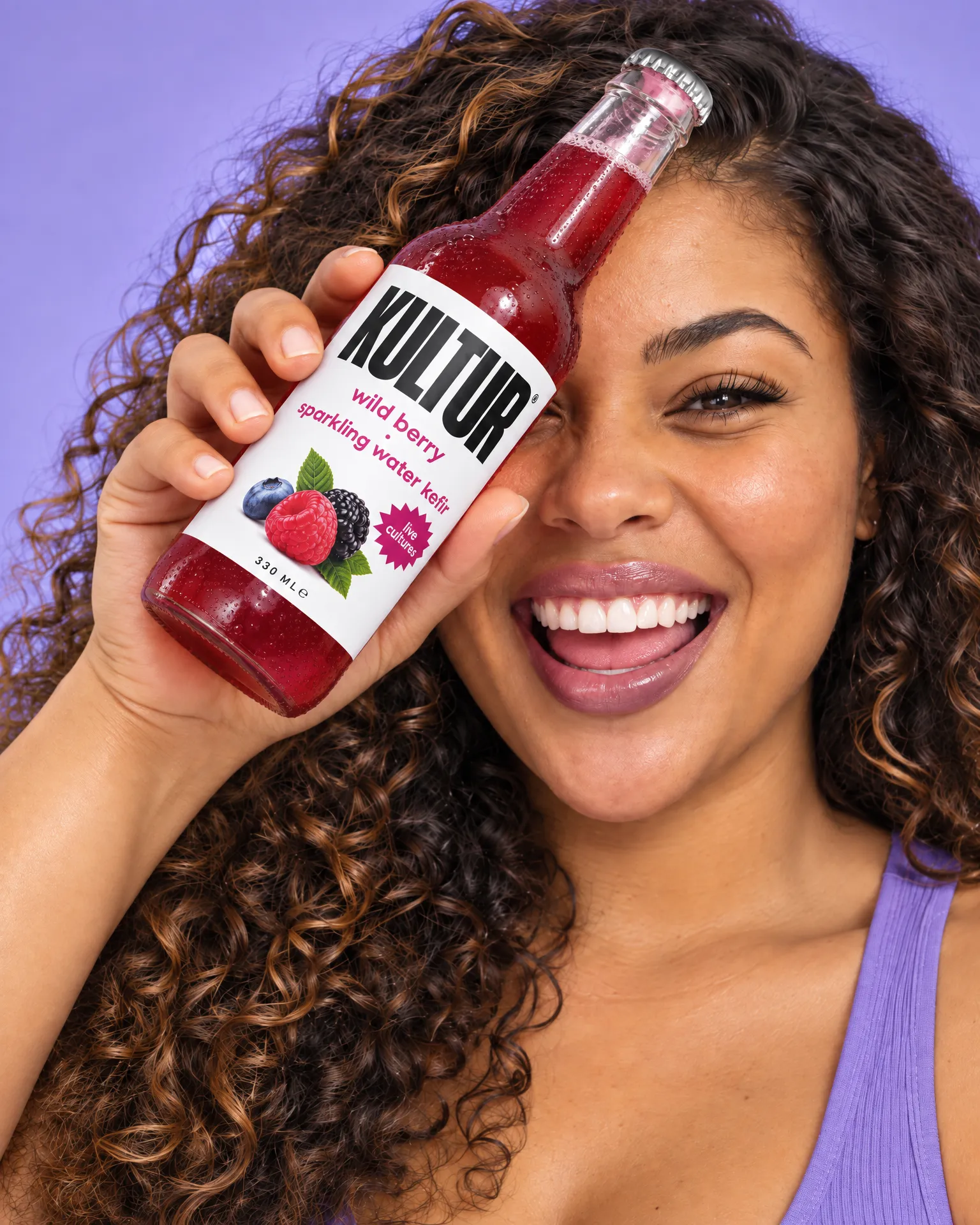 Luz laughs, a bottle of Kultur sparkling kefir held beside her eye, on lilac.