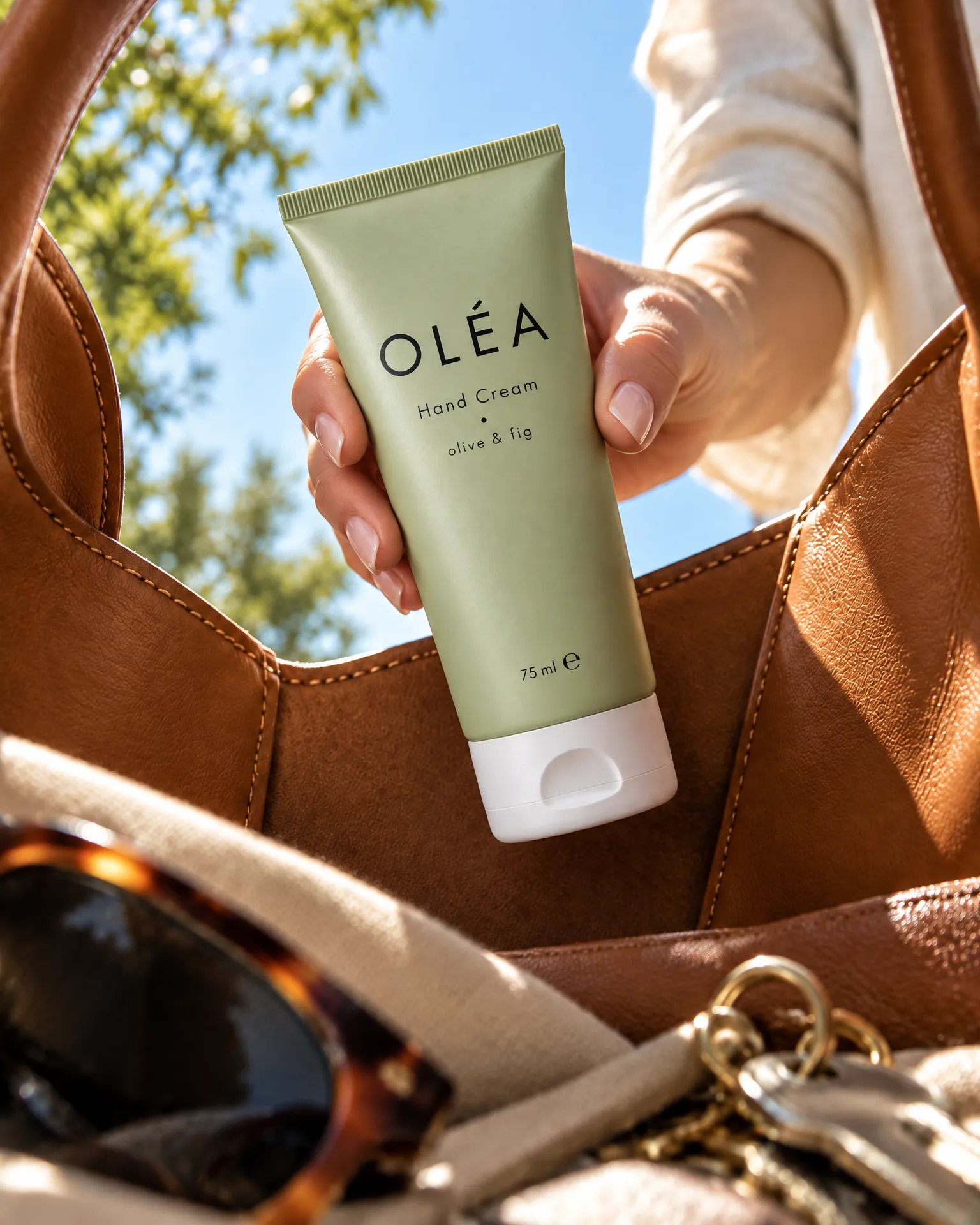 A hand lifts a tube of Oléa hand cream out of a tan leather tote, seen from inside the bag.