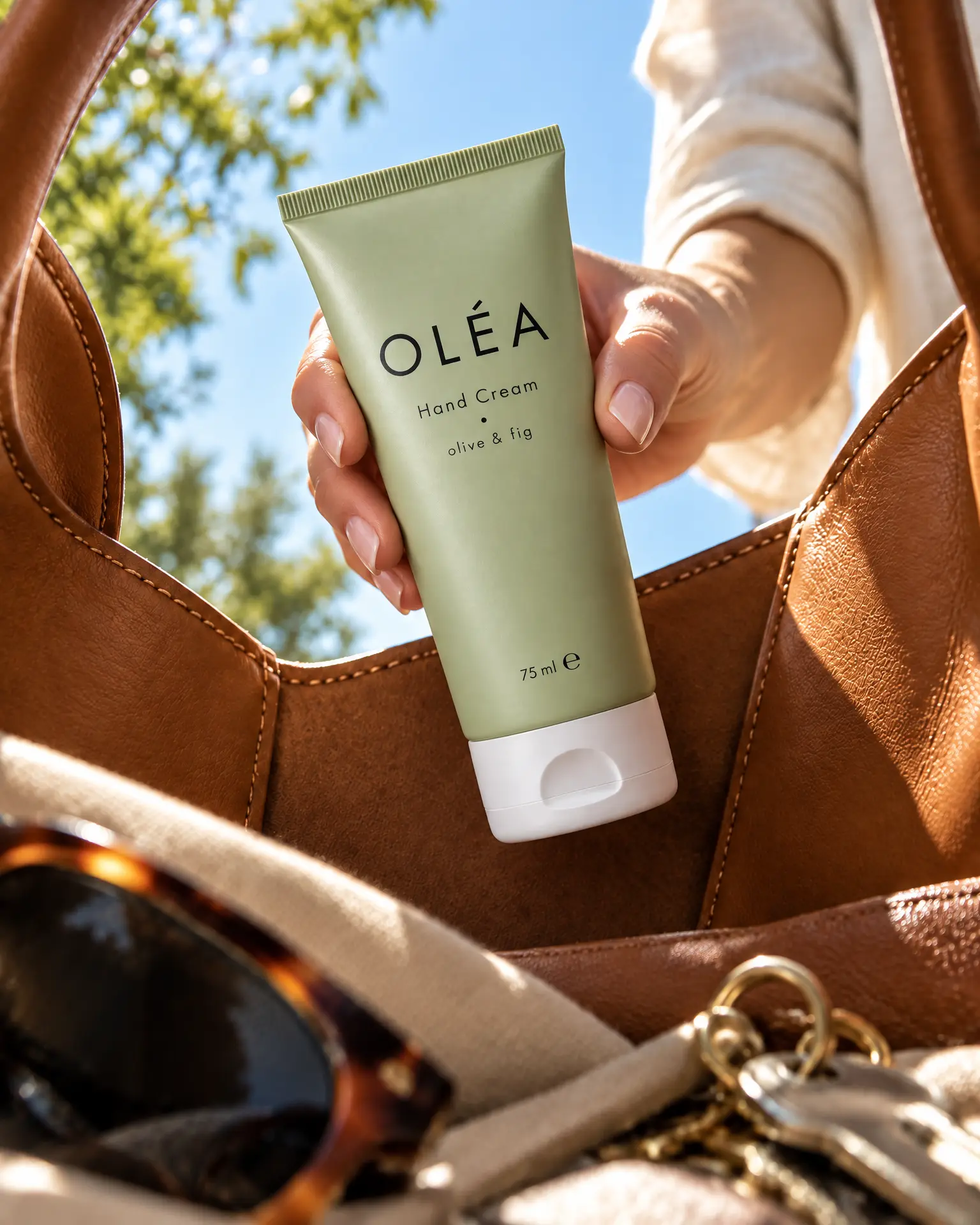 A hand lifts a tube of Oléa hand cream out of a tan leather tote, seen from inside the bag.