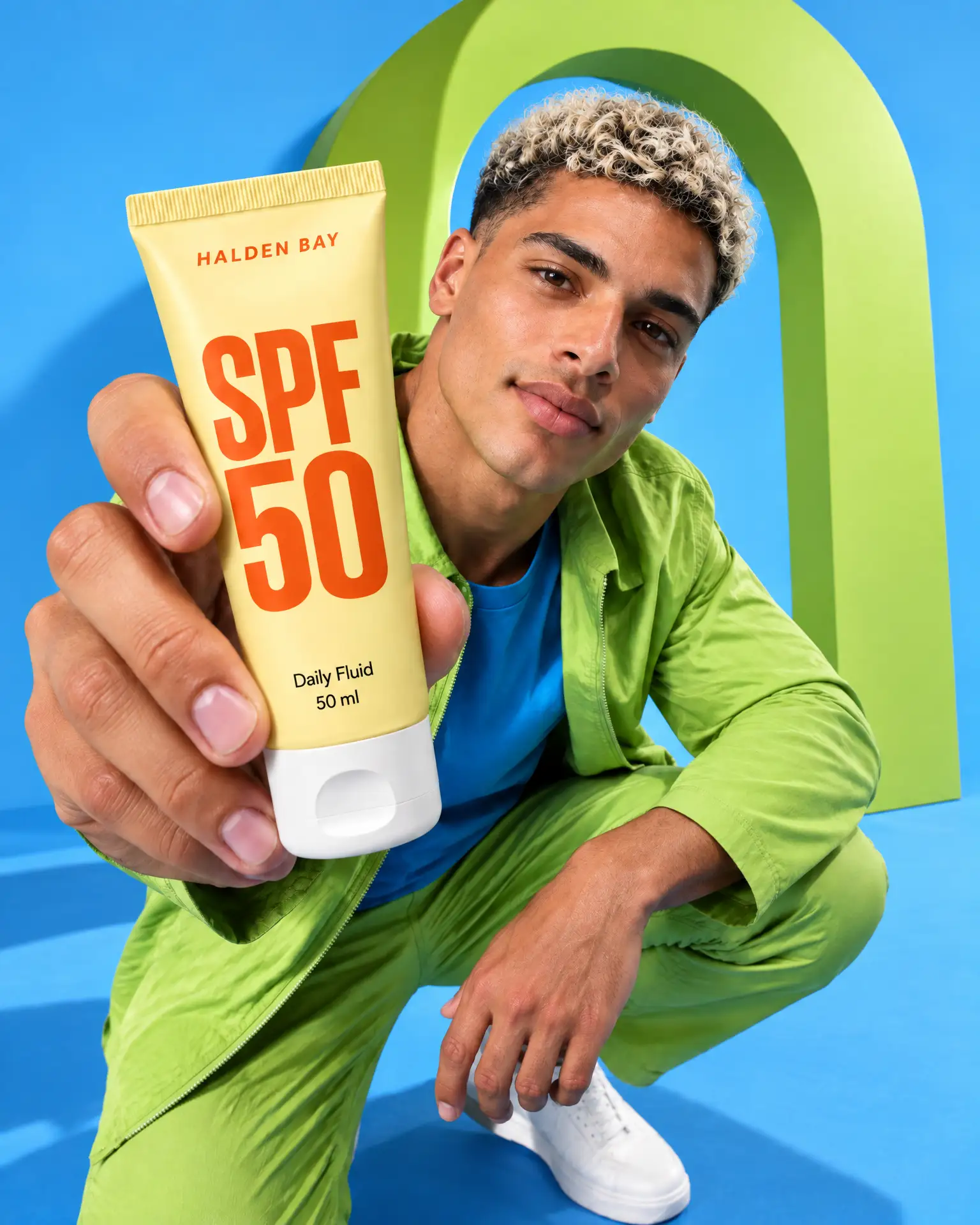 Nico in lime green crouches under a lime arch on sky blue, holding a yellow SPF 50 tube out to the lens.