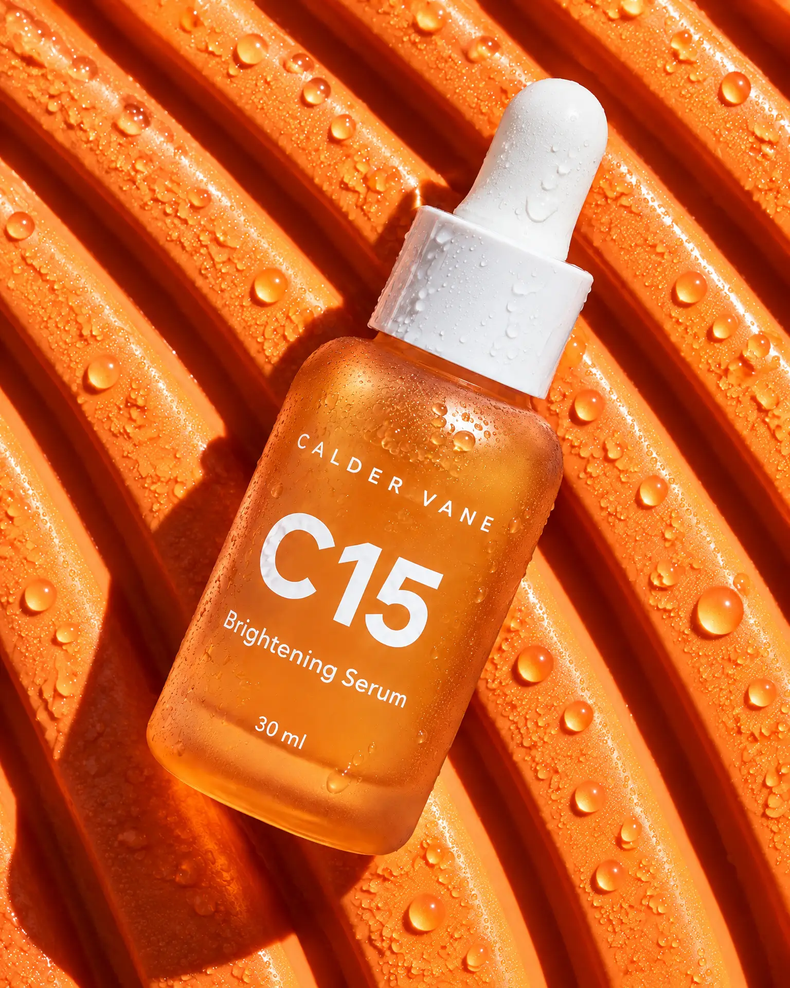 A frosted orange Calder Vane C15 serum bottle lying on wet orange ribbed rubber, seen from above.