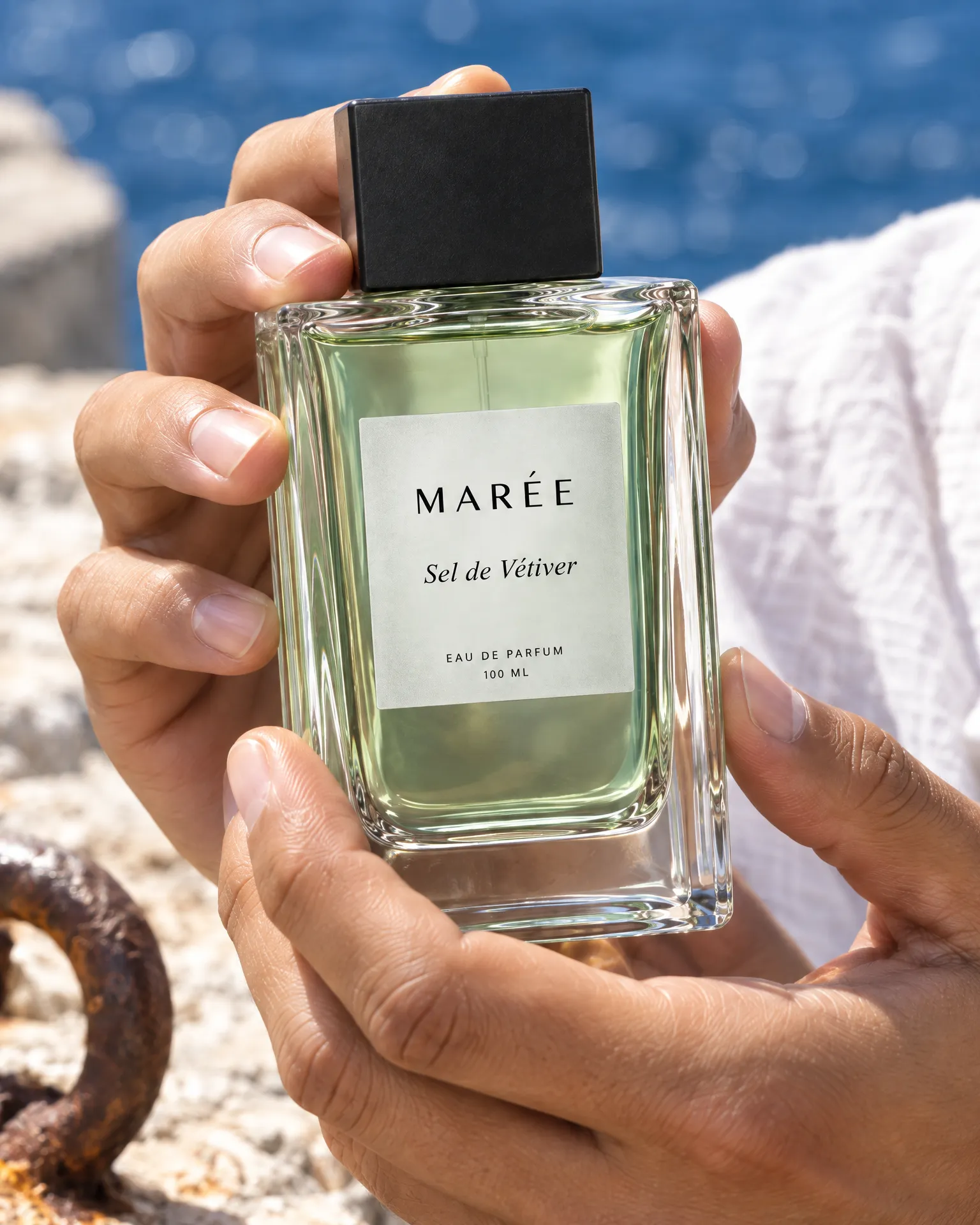 Nico's hands holding a bottle of Marée Sel de Vétiver close to the lens against the sea wall's concrete, the sea soft behind.