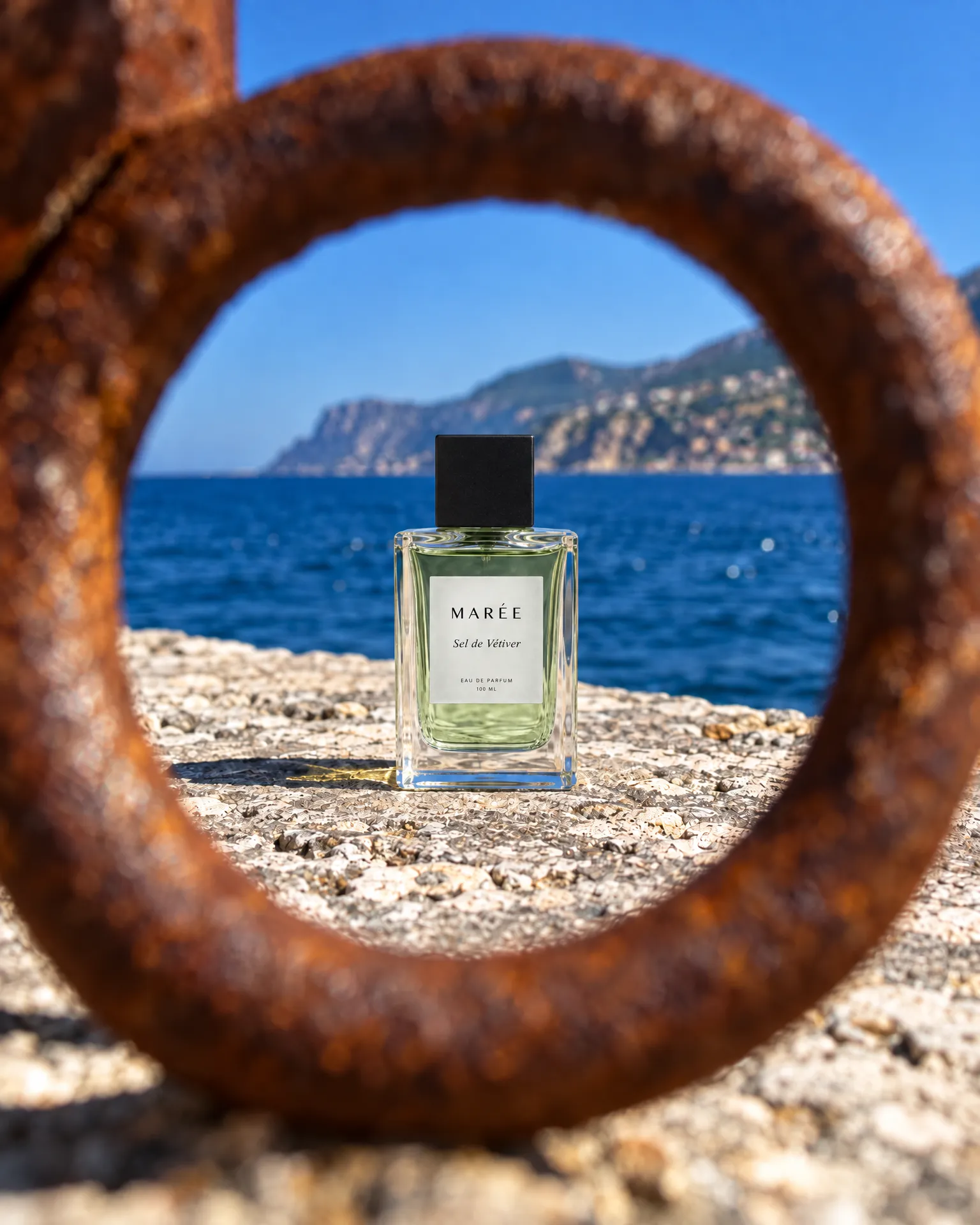 A bottle of Marée Sel de Vétiver standing on a sea wall, framed by a rusted iron mooring ring in the foreground, the blue sea and a headland behind.